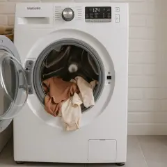 Lavadora Samsung con ropa de bebé lista para lavar. La puerta abierta muestra varios bodies de algodón en tonos tierra y pastel.