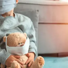 Niño con mascarilla abraza a su osito, también con mascarilla. Una imagen que refleja la realidad del **covid en bebes** y niños durante la pandemia.
