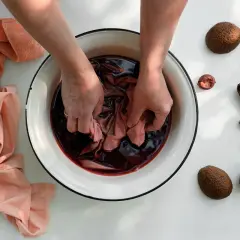 Manos sumergiendo tela en tinte oscuro para aprender como teñir ropa a mano. Cáscaras de aguacate alrededor.
