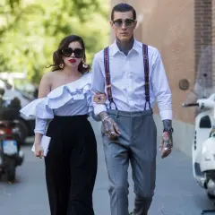 Pareja elegante camina, él es un invitado boda hombre sin traje, con tirantes y tatuajes visibles.