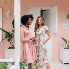 Dos mujeres posan frente a una casa rosa. Una lleva un vestido rosa liso, la otra uno floral. Ideas para como ir a una boda informal mujer.