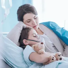 Niña con mascarilla de oxígeno y oso de peluche, su madre la abraza. Posibles síntomas de neumonía en niños.