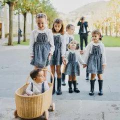 Niños de arras, con vestidos de cuadros y botas, pasean sonrientes. Un bebé en una cesta de mimbre les acompaña.
