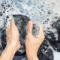 Manos lavando ropa oscura en una tina con espuma. Se puede lavar la ropa con gel de baño para una limpieza suave.
