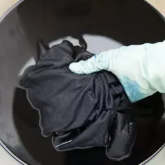 Mano con guante sumerge tela negra en líquido oscuro para recuperar color de ropa oscura.