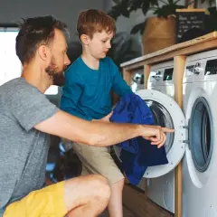 Padre e hijo aprenden como lavar ropa de deporte. El niño ayuda a meter la ropa azul en la lavadora.