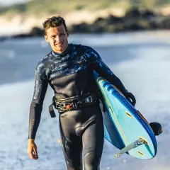 Hombre sonriente con tabla de kitesurf en la playa. Su traje de neopreno es impermeable, ideal para deportes acuáticos.
