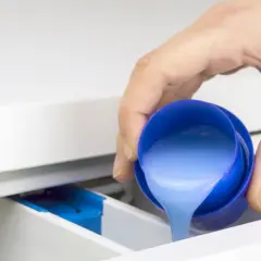 Mano vierte líquido azul, que parece suavizante y detergente, en el cajetín de la lavadora.