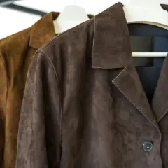 Dos chaquetas de ante, una marrón y otra café, colgadas en perchas. ¿Sabías que se puede lavar una chaqueta de ante en la lavadora?