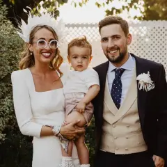 Familia feliz en una boda. La novia, con un tocado blanco y gafas redondas, abraza a su hijo. El novio, con traje oscuro y chaleco beige, sonríe. Son los pajes boda más adorables.