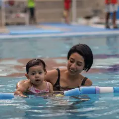 Madre sonriente enseña a su bebé natación para niños en la piscina, rodeados de flotadores.