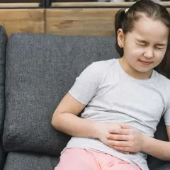 Niña con dolor de barriga en niños cuando preocuparse. Se agarra el abdomen con expresión de malestar sentada en un sofá.