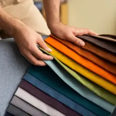 Manos sostienen muestras de tela de poliéster en tonos vibrantes y neutros, sobre un sillón gris.