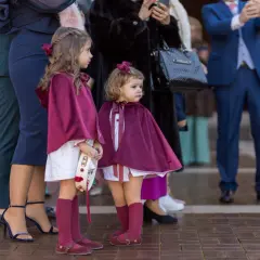 Dos niñas con capas de terciopelo borgoña, vestidas para una boda de invierno, esperan junto a adultos elegantes.