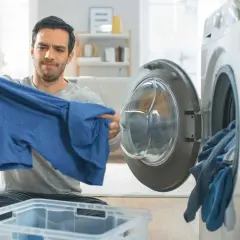 Hombre preocupado revisa una camiseta azul, pensando en como lavar algodón para que no encoja. Cerca, una lavadora abierta con ropa.