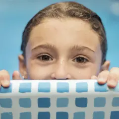 Niña con ojos curiosos se aferra al borde de la piscina, lista para aprender como enseñar a nadar a un niño.
