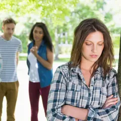 Una chica se siente sola y triste, apoyada en un árbol, mientras sus amigos hablan en el fondo. Es un momento típico de cuando empieza la adolescencia.