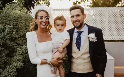 Familia feliz en una boda. La novia, con un tocado blanco y gafas redondas, abraza a su hijo. El novio, con traje oscuro y chaleco beige, sonríe. Son los pajes boda más adorables.