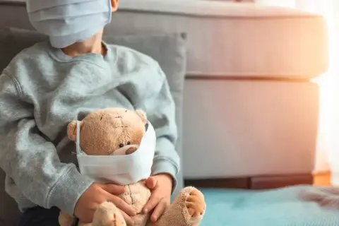 Niño con mascarilla abraza a su osito, también con mascarilla. Una imagen que refleja la realidad del **covid en bebes** y niños durante la pandemia.