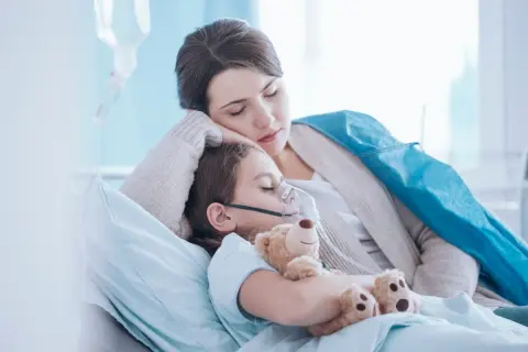 Niña con mascarilla de oxígeno y oso de peluche, su madre la abraza. Posibles síntomas de neumonía en niños.