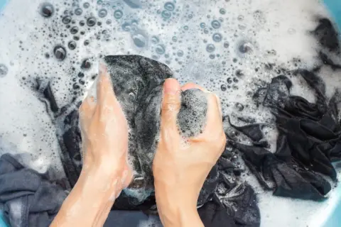 Manos lavando ropa oscura en una tina con espuma. Se puede lavar la ropa con gel de baño para una limpieza suave.