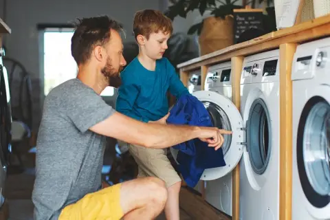 Padre e hijo aprenden como lavar ropa de deporte. El niño ayuda a meter la ropa azul en la lavadora.