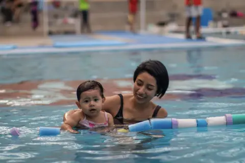 Madre sonriente enseña a su bebé natación para niños en la piscina, rodeados de flotadores.