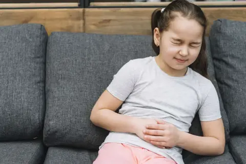 Niña con dolor de barriga en niños cuando preocuparse. Se agarra el abdomen con expresión de malestar sentada en un sofá.