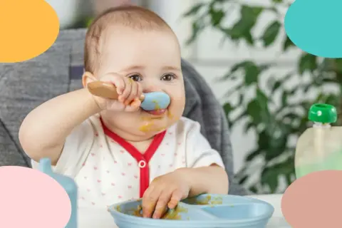 Un bebé de 12 meses, con la cara manchada de comida, intenta alimentarse solo con una cuchara azul.