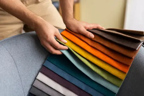 Manos sostienen muestras de tela de poliéster en tonos vibrantes y neutros, sobre un sillón gris.