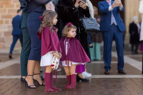 Dos niñas con capas de terciopelo borgoña, vestidas para una boda de invierno, esperan junto a adultos elegantes.