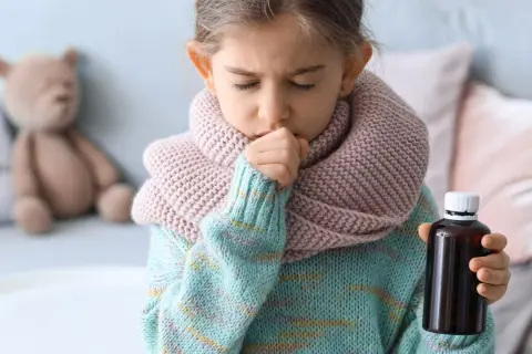 Niña con tos y bufanda, sosteniendo un frasco de medicina. Una imagen que ilustra cómo calmar la tos en niños.