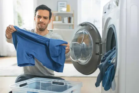 Hombre preocupado revisa una camiseta azul, pensando en como lavar algodón para que no encoja. Cerca, una lavadora abierta con ropa.