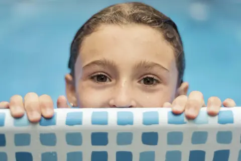 Niña con ojos curiosos se aferra al borde de la piscina, lista para aprender como enseñar a nadar a un niño.