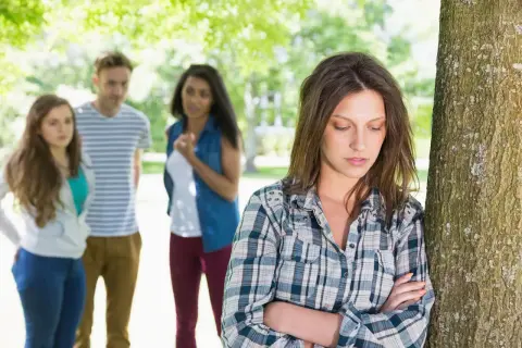 Una chica se siente sola y triste, apoyada en un árbol, mientras sus amigos hablan en el fondo. Es un momento típico de cuando empieza la adolescencia.