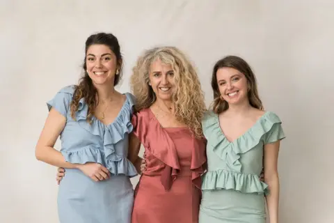 Tres mujeres sonríen, vestidas con trajes elegantes. Ideas para como vestir para un bautizo en invierno.