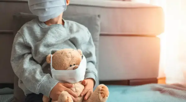 Niño con mascarilla abraza a su osito, también con mascarilla. Una imagen que refleja la realidad del **covid en bebes** y niños durante la pandemia.