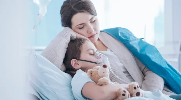 Niña con mascarilla de oxígeno y oso de peluche, su madre la abraza. Posibles síntomas de neumonía en niños.
