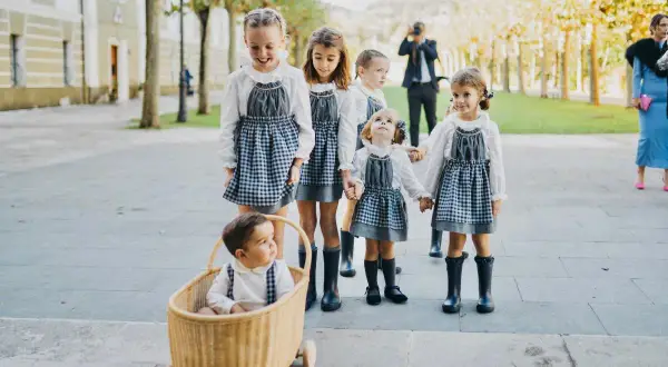 Niños de arras, con vestidos de cuadros y botas, pasean sonrientes. Un bebé en una cesta de mimbre les acompaña.