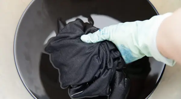 Mano con guante sumerge tela negra en líquido oscuro para recuperar color de ropa oscura.