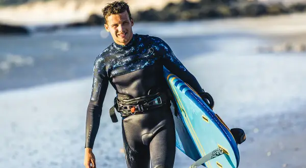 Hombre sonriente con tabla de kitesurf en la playa. Su traje de neopreno es impermeable, ideal para deportes acuáticos.