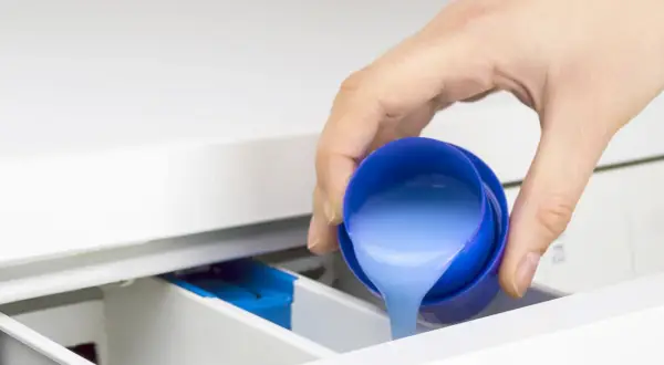 Mano vierte líquido azul, que parece suavizante y detergente, en el cajetín de la lavadora.