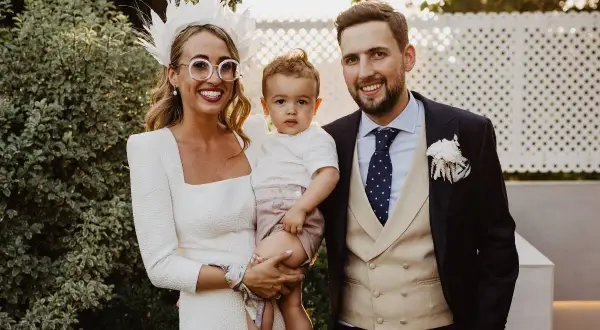 Familia feliz en una boda. La novia, con un tocado blanco y gafas redondas, abraza a su hijo. El novio, con traje oscuro y chaleco beige, sonríe. Son los pajes boda más adorables.
