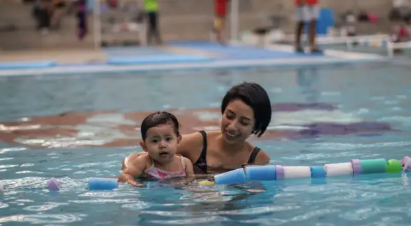 Madre sonriente enseña a su bebé natación para niños en la piscina, rodeados de flotadores.