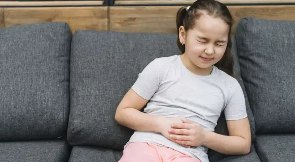 Niña con dolor de barriga en niños cuando preocuparse. Se agarra el abdomen con expresión de malestar sentada en un sofá.