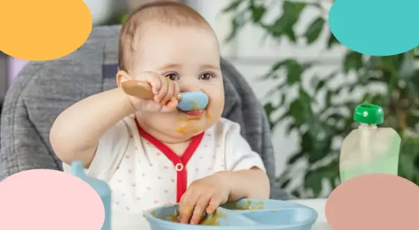Un bebé de 12 meses, con la cara manchada de comida, intenta alimentarse solo con una cuchara azul.