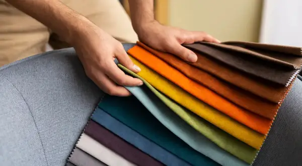 Manos sostienen muestras de tela de poliéster en tonos vibrantes y neutros, sobre un sillón gris.