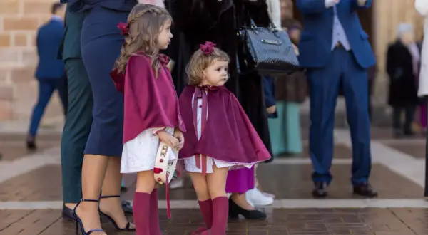 Dos niñas con capas de terciopelo borgoña, vestidas para una boda de invierno, esperan junto a adultos elegantes.