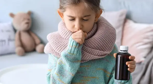 Niña con tos y bufanda, sosteniendo un frasco de medicina. Una imagen que ilustra cómo calmar la tos en niños.
