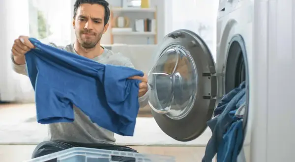 Hombre preocupado revisa una camiseta azul, pensando en como lavar algodón para que no encoja. Cerca, una lavadora abierta con ropa.